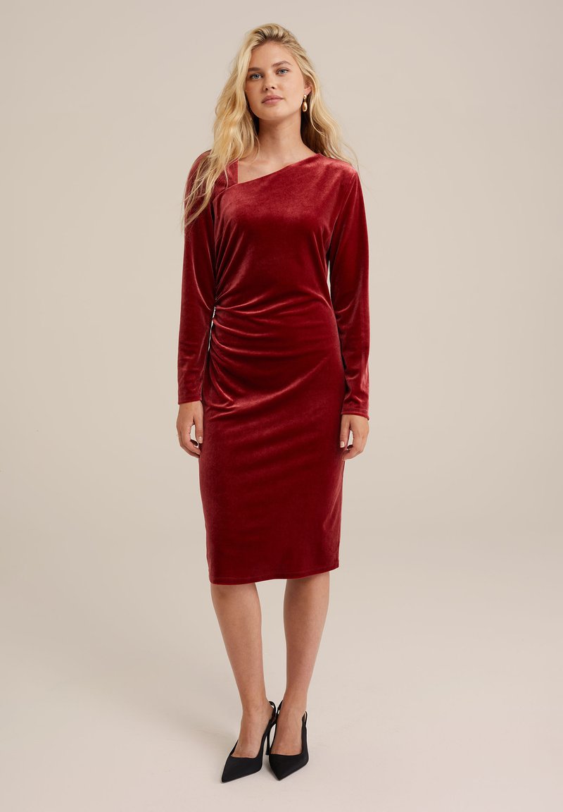 Czerwona sukienka z welwetu z długimi rękawami, asymetrycznym dekoltem, dopasowanym krojem, marszczeniem po boku i długością do kolan. Czarne szpilki.