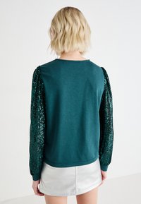 Sudadera en color verde azulado con mangas de puff cubiertas de lentejuelas, escote redondo y un ajuste holgado, combinada con una falda corta plateada que presenta un patrón texturizado.