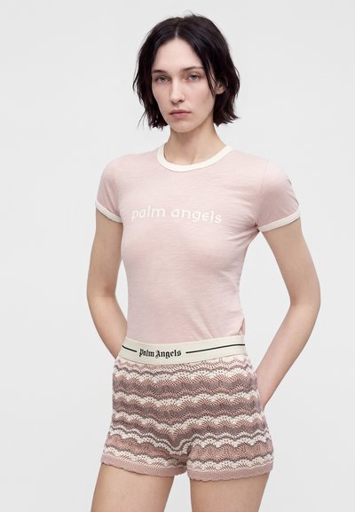 Νεαρή γυναίκα που φοράει ένα ανοιχτό ροζ t-shirt "palm angels" και πλεκτές σορτς με σχέδιο και branded ελαστική μέση.