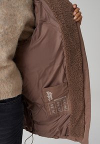 Veste isolée marron avec une doublure intérieure en peluche. Elle dispose d'une poche sécurisée, d'un tissu lisse et d'une texture agréable autour du col.