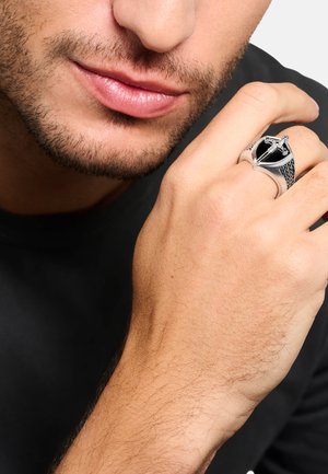 Nahaufnahme des unteren Gesichts und der Hand eines Mannes, der einen silbernen Ring mit einem Schwertdesign auf seinem Finger vor einem schwarzen Hemdenhintergrund trägt.