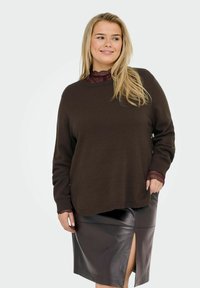 Femme souriante avec de longs cheveux blonds, portant un pull marron superposé à un top en dentelle et une jupe en cuir marron avec une fente à l'avant.