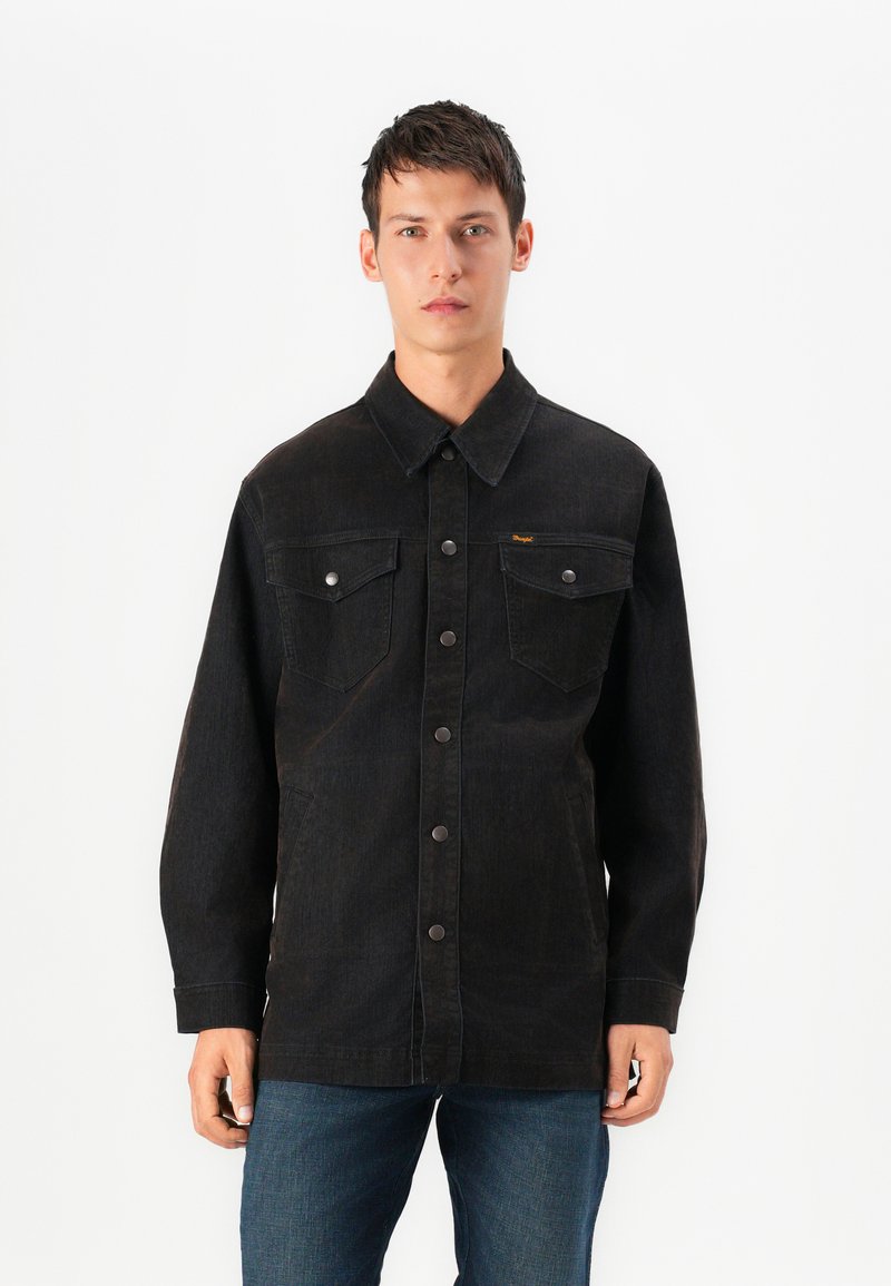 Črna denim jakna s ovratnikom, sprednjim gumbom, dvema žepoma na prsih in subtilnimi šivi. Nosi se čez modre kavbojke.