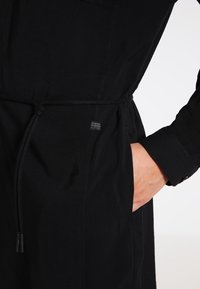 Vestido camisa ligero de color negro con cinturón ajustable en la cintura, bolsillos laterales y etiqueta de tela. Presenta un puño abotonado y una textura suave.