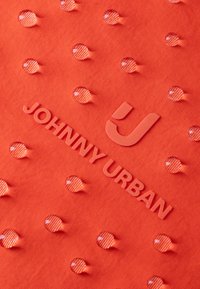 Piros szövet texturált felülettel, kiemelkedő cseppekkel és dombornyomott "JOHNNY URBAN" logóval, sima, kontrasztszínű elemekkel.