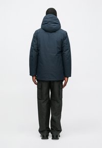 Muž oblečený v tmavomodrej kapucňovej bunde, čiernych cargo nohaviciach s bočnými vreckami a čiernych topánkach, stojaci otočený chrbtom k svetlému pozadiu.