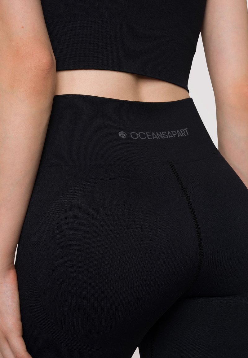 Leggings neri a vita alta realizzati in un tessuto morbido e elastico. Presentano una cintura elastica con il logo "OCEANSAPART" e dettagli di cucitura.