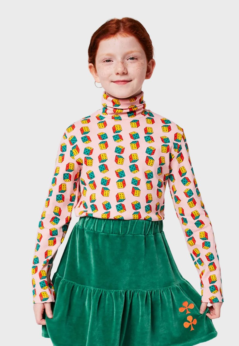 Langärmliges pinkes Oberteil mit einem wiederholten Muster aus bunten Würfeln; kombiniert mit einem grünen Samt-Tüllrock mit orangefarbener Blumenstickerei.
