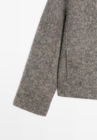 Manica di un cappotto grigio di lana con finitura testurizzata, caratterizzata da un taglio dritto, una tasca singola e dettagli di cucitura visibili al polsino.