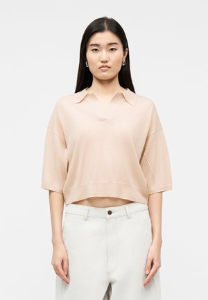 Fiatal nő hosszú sötét hajjal, laza, halványpink rövid ujjú inget és magas derekú fehér nadrágot visel, egyszerű fehér háttér előtt állva.