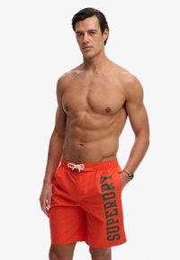 Homme torse nu aux cheveux courts et foncés, portant un short de bain orange avec le texte "SUPERDRY" et une montre-bracelet, debout devant un fond uni.