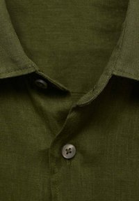 Colletto di camicia verde oliva in tessuto texturizzato, con chiusura a bottone dotata di quattro fori. Linee pulite e semplici in tutto.