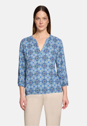 Vrouw draagt een blauw, langärmelig blouse met patroon en V-hals, gecombineerd met lichtbeige broek, staand tegen een effen achtergrond.