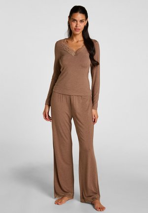 Femme debout pieds nus portant un haut marron à manches longues et un pantalon à jambes larges avec des détails en dentelle sur l'encolure et les ourlets du pantalon.