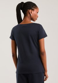 Moteris su įrišta kasytė, vilkinti juodą trumpomis rankovėmis paprastą marškinėlį ir juodas kelnes, žiūrinti nuo nugaros priešais paprastą foną.