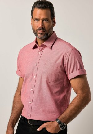 Man met baard die een roze overhemd met korte mouwen en knopen draagt, met opgerolde mouwen en een polshorloge, staand tegen een effen lichte achtergrond.