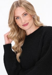 Černý pletený svetr s texturovaným vzorem a širokými rukávy, doplněný o vrstvené blond vlasy a přirozený make-up, který prezentuje neutrální vzhled.