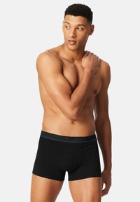 Černé boxerské trenýrky s jemným diamantovým vzorem, elastickým páskem a hladkou texturou. Model prezentuje trenýrky v uvolněné póze.