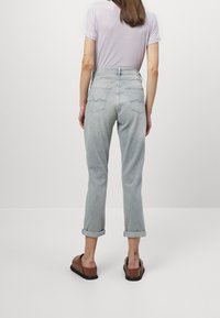 Un jean en denim gris clair présente une taille haute, des ourlets retroussés et deux poches arrière avec des coutures courbées. Porté avec un haut léger simple et des sandales marron.