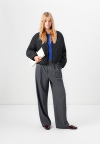 Jeune femme avec de longs cheveux blonds, portant une veste matelassée noire, un pull bleu et un pantalon large gris, tenant une pochette blanche sur fond blanc.