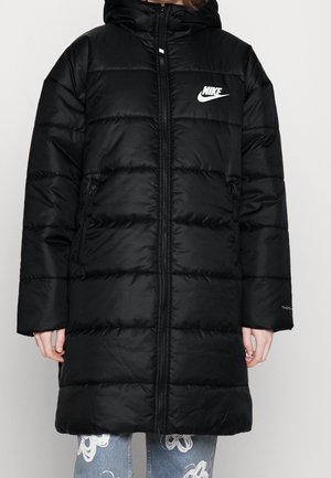 Manteau à capuche matelassé noir Nike avec fermeture éclair frontale, poches zippées et logo blanc sur la poitrine, porté sur un jean bleu avec des motifs blancs.