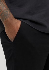 Zwarte stoffen broek met een zijzak. Glad textuur, perfecte pasvorm en een close-up weergave van een arm die in de zak rust.