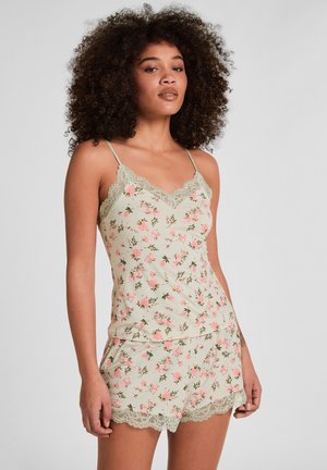 Frau mit lockigem Haar, die ein blassgrünes, florales Camisole und passende Shorts mit Spitzenbesatz trägt, stehend vor einfachem Hintergrund.