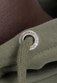 Olíva zöld szövet fonott csipkével, melyen egy fém gyűrű található, amelyen az "ALPHA INDUSTRIES USA" felirat látható, és textúrált barna háttér jellemzi.
