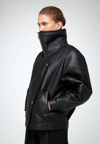 Zwarte leren jas met een hoge kraag en een asymmetrische rits. Heeft een oversized pasvorm, zijzakken en minimale hardwaredetails.