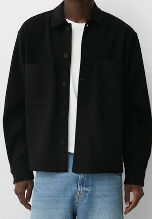 Schwarze Wollhemdjacke mit Kragen, Frontknopfverschluss, zwei großen Brusttaschen und langen Ärmeln, kombiniert mit hellblauen Jeans.