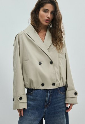 Femme portant une veste courte beige à double boutonnage avec de grands revers et des boutons foncés, associée à un jean en denim bleu.