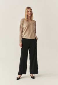Beige sveter s výstrihom do V vyrobený z jemného pleteného materiálu, skombinovaný s vysokosťahovanými čiernymi nohavicami s širokými nohavicami. Modelka má na sebe čierne lodičky s ostrou špičkou.