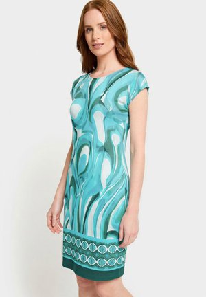Woman wearing a sleeveless knee-length dress with abstract blue, teal, and white swirl patterns and a geometric border near the hem.