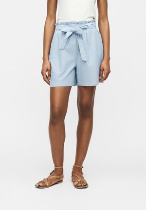Kvinne iført lyseblå høytaljede shorts med stoffbelte knytt i sløyfe, kombinert med brune flate sandaler og hvit ermeløs topp.