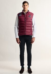 Chaleco de plumas burdeos con cierre de cremallera, costuras horizontales y parches. Lleva encima una camisa blanca, combinado con pantalones negros y zapatos marrones.