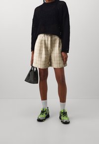 Czarny luźny sweter dziany; beżowe teksturowane szorty; czarna siatkowa torba; zielono-szare buty sportowe; białe skarpetki sięgające do kostek.