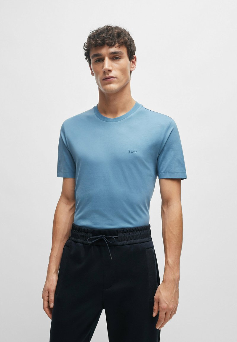 T-shirt azzurro chiaro a maniche corte realizzato in tessuto morbido; presenta un colletto a girocollo e un discreto dettaglio del logo sul petto. Indossato con pantaloni neri.