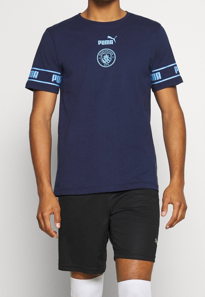 Man med en marinblå Manchester City Puma-t-shirt med logotyp på bröstet och ärmarna, tillsammans med svarta Puma-shorts och vita strumpor.