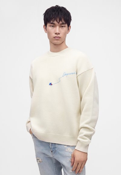 Jeune homme portant un pull crème avec de petites broderies bleues et rouges et un jean bleu clair déchiré, debout devant un fond uni.