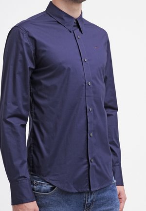 Mann trägt ein eng anliegendes dunkelblaues Hemd mit langen Ärmeln und Knöpfen, mit kleinem Logo auf der Brust und blaue Jeans.