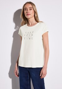 Lysegult kortærmet t-shirt med rund halsudskæring, der har teksten "TAKE YOUR TIME" i mørkegrå, parret med mørkeblå jeans.