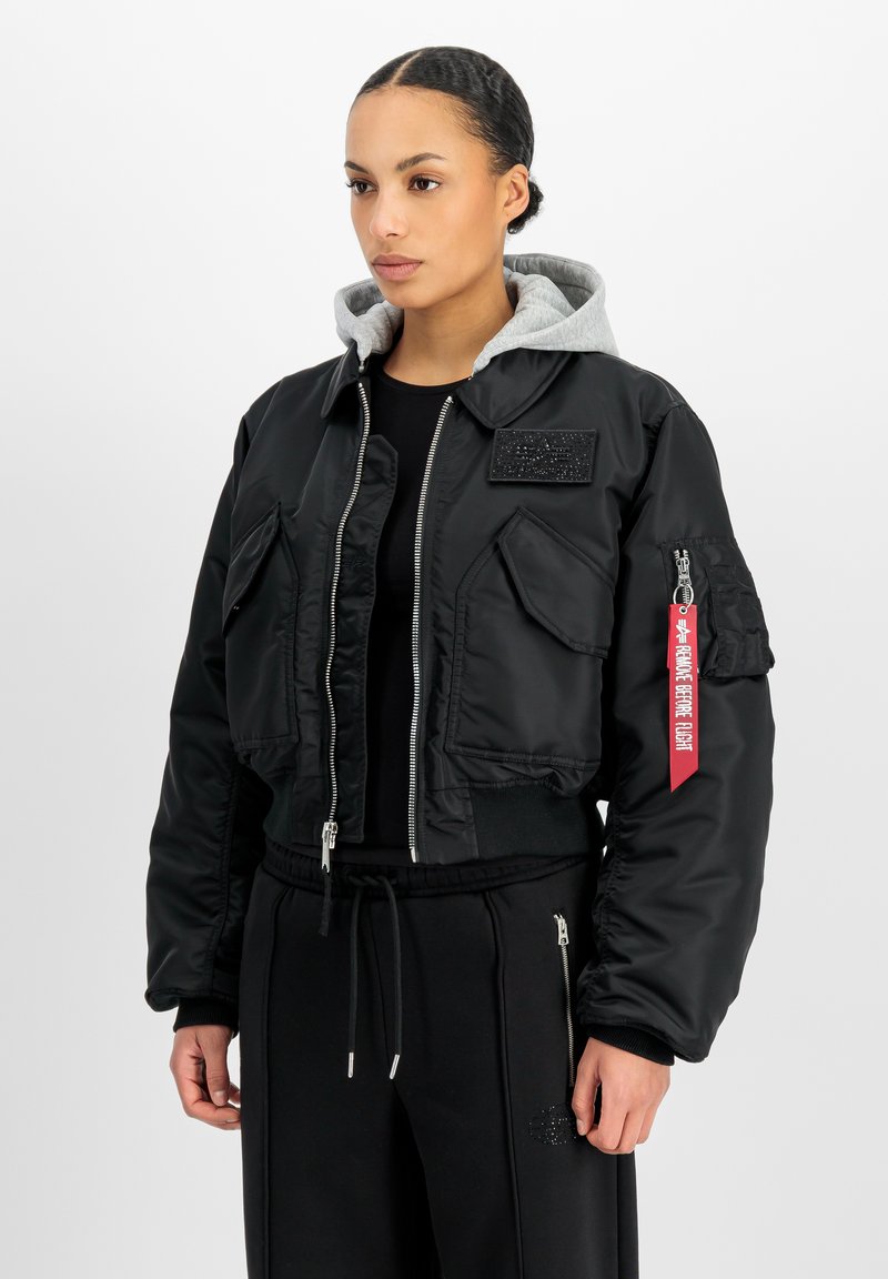 Γυναίκα που φοράει μαύρο bomber jacket με μεγάλες τσέπες, γκρι φούτερ με κουκούλα από μέσα και μαύρο παντελόνι με κορδόνια.