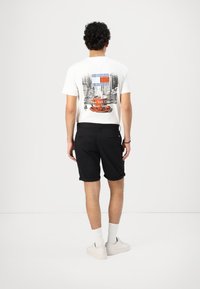 T-shirt blanc avec un imprimé graphique représentant un paysage urbain et du texte au dos. Un short noir et des baskets claires complètent la tenue.