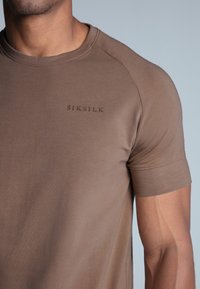 Smeđi kratki rukav T-shirt napravljen od glatkog materijala, s okruglim izrezom i utisnutim logotipom "SIKSILK" na lijevoj strani prsnog koša.