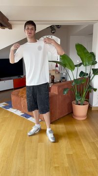 Ung mann innendørs som holder opp en hvit T-skjorte med en liten spiral logo, iført svarte shorts og sølv sneakers, nær en stor potteplante.