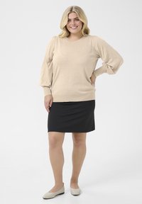 Pull en tricot beige à manches bouffantes, assorti à une jupe noire jusqu'aux genoux et des ballerines beiges, positionné sur un fond gris clair.