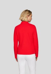 Red, textured pullover with a high collar and long sleeves, featuring a fitted design and subtle seam details, worn with white pants.
