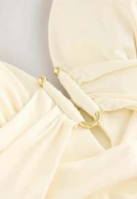 Crème-kleurige stof met een gladde textuur, voorzien van een gouden accentring en geplooide details aan de randen. Royaal, zachte cups.