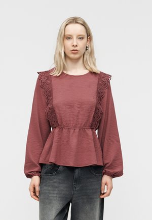 Γυναίκα με μακρυμάνικο πουκάμισο σε αποχρώσεις του μωβ με δαντελένιες λεπτομέρειες στους ώμους και ζώνη peplum, συνδυασμένο με ξεθωριασμένο μαύρο τζιν.