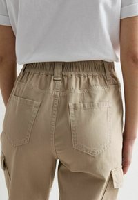 Pantaloni cargo beige con vita elastica, dotati di due tasche posteriori e cuciture in vista. Il tessuto sembra resistente e testurizzato.